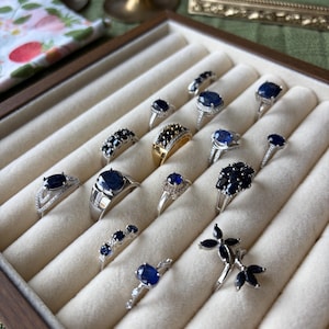 May include: A collection of silver and gold rings with dark blue gemstones displayed in a wooden jewelry box. The rings feature various designs, including solitaire, cluster, and band styles. The gemstones are oval, round, and marquise cuts.