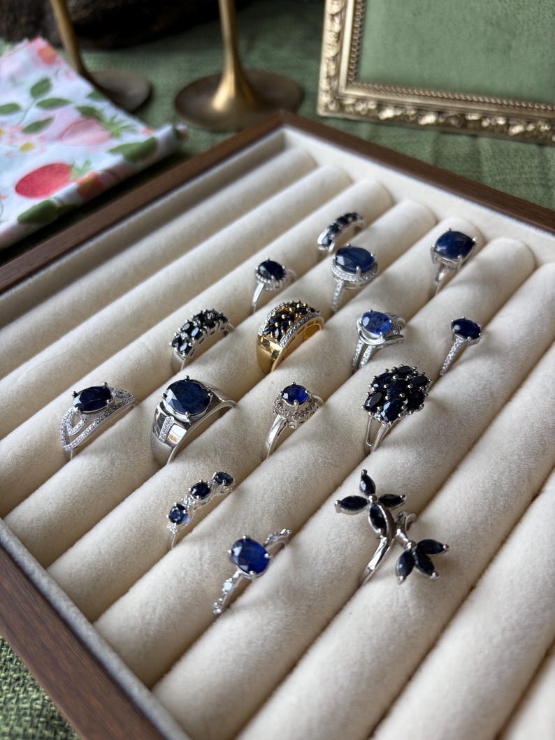 May include: A collection of silver and gold rings with dark blue gemstones displayed in a wooden jewelry box. The rings feature various designs, including solitaire, cluster, and band styles. The gemstones are oval, round, and marquise cuts.