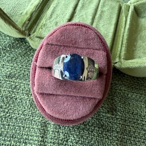 May include: A silver ring with a large, oval, dark blue gemstone, possibly a sapphire, set in a band with small accent stones. The ring rests on a pink velvet ring box, with a green velvet jewelry case in the background.