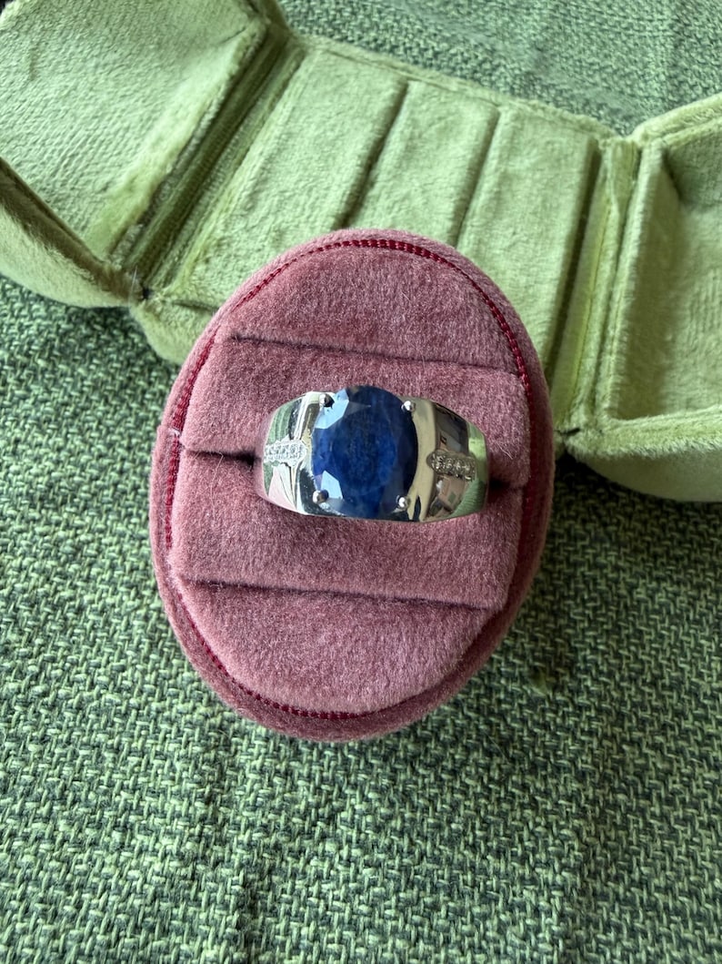 May include: A silver ring with a large, oval, dark blue gemstone, possibly a sapphire, set in a band with small accent stones. The ring rests on a pink velvet ring box, with a green velvet jewelry case in the background.