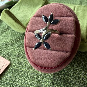May include: A silver ring with a floral design, featuring black gemstone petals. The ring is displayed on a pink ring box with a red trim, set against a green textured background.