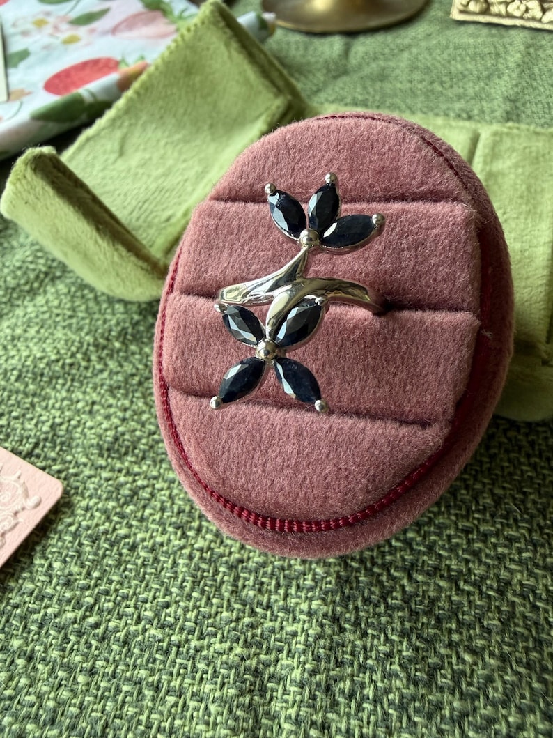 May include: A silver ring with a floral design, featuring black gemstone petals. The ring is displayed on a pink ring box with a red trim, set against a green textured background.