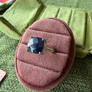 May include: A silver ring with a large, square, dark blue gemstone set in a prong setting. The ring is displayed in a pink velvet ring box with a green velvet background.