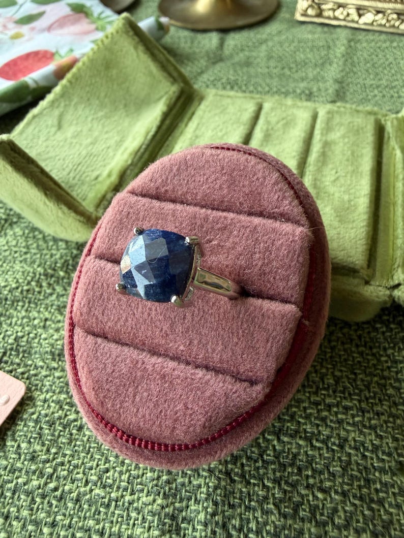 May include: A silver ring with a large, square, dark blue gemstone set in a prong setting. The ring is displayed in a pink velvet ring box with a green velvet background.
