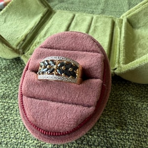 May include: A gold ring with a rectangular face, set with dark blue gemstones and small clear stones. The ring is displayed in a pink velvet ring box with a red trim, inside a green velvet jewelry box.