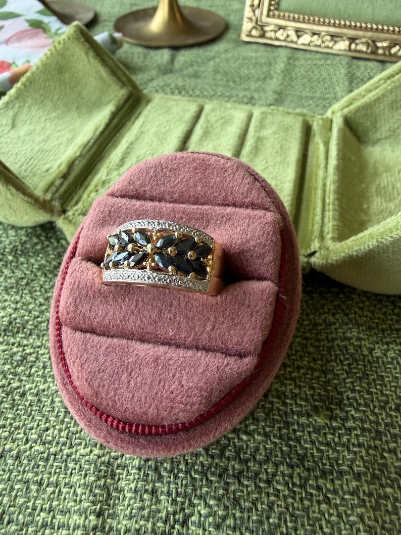 May include: A gold ring with a rectangular face, set with dark blue gemstones and small clear stones. The ring is displayed in a pink velvet ring box with a red trim, inside a green velvet jewelry box.
