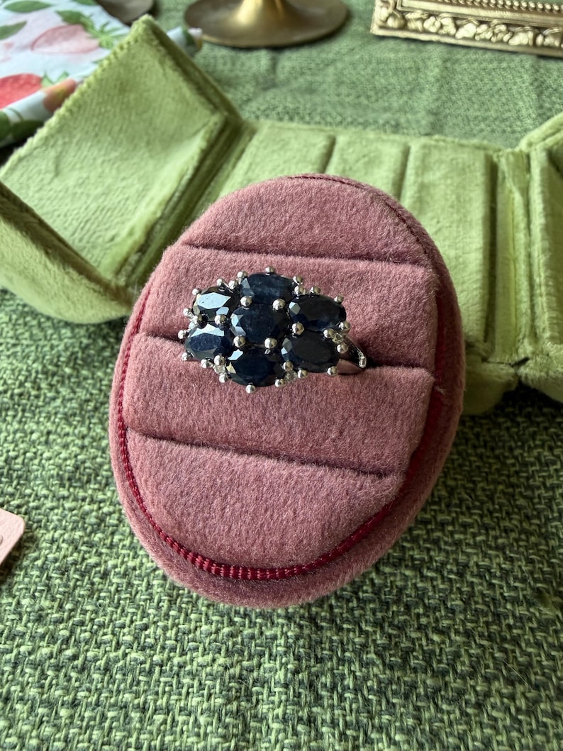 May include: A silver ring with a cluster of dark blue oval gemstones set on a pink velvet ring box. The ring box is inside a green velvet jewelry case, set on a green textured surface.