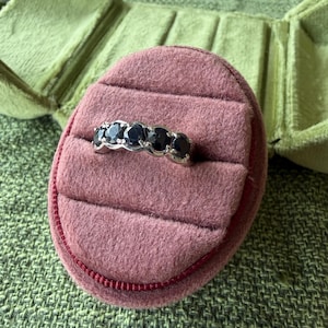 May include: A silver ring with five dark blue gemstones set in a row. The ring is displayed on a pink velvet ring box, with a green velvet jewelry box in the background. The ring box is on a green textured surface.