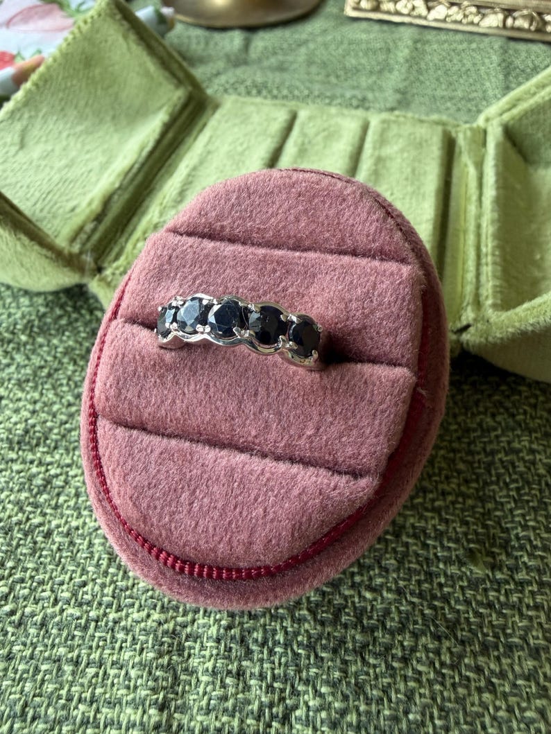 May include: A silver ring with five dark blue gemstones set in a row. The ring is displayed on a pink velvet ring box, with a green velvet jewelry box in the background. The ring box is on a green textured surface.