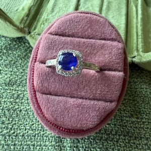 May include: A silver ring with a large, oval, deep blue gemstone surrounded by a halo of smaller, clear stones. The ring is set on a pink velvet ring box, with a green fabric background.