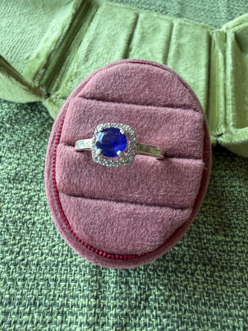 May include: A silver ring with a large, oval, deep blue gemstone surrounded by a halo of smaller, clear stones. The ring is set on a pink velvet ring box, with a green fabric background.