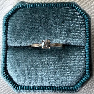 May include: A gold ring with a single, round, clear gemstone is displayed inside a teal velvet ring box. The ring box is octagonal and has a teal border. The background is a light gray fabric.