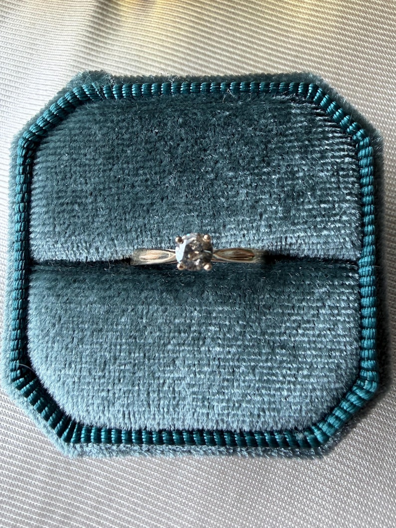 May include: A gold ring with a single, round, clear gemstone is displayed inside a teal velvet ring box. The ring box is octagonal and has a teal border. The background is a light gray fabric.
