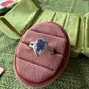 May include: A silver ring with a large, oval, blue gemstone is displayed on a pink velvet ring box. The ring has a split band and is accented with smaller, clear gemstones. The ring box is inside a green velvet jewelry case.