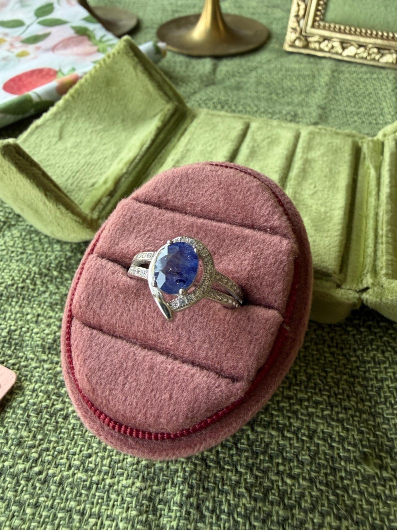 May include: A silver ring with a large, oval, blue gemstone is displayed on a pink velvet ring box. The ring has a split band and is accented with smaller, clear gemstones. The ring box is inside a green velvet jewelry case.