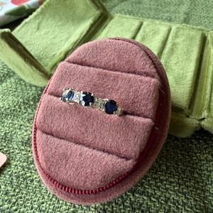 May include: A silver ring with three dark blue gemstones and small clear stones, set in a pink velvet ring box. The ring box is open, revealing the ring against a green background.