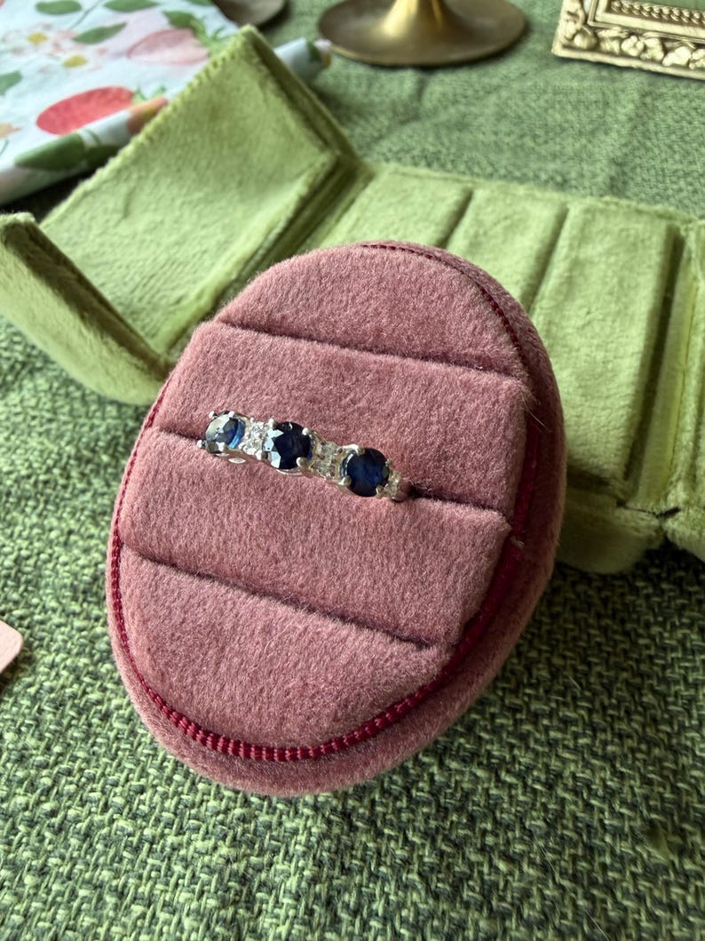 May include: A silver ring with three dark blue gemstones and small clear stones, set in a pink velvet ring box. The ring box is open, revealing the ring against a green background.