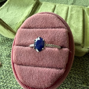 May include: A silver ring with a large, oval, dark blue gemstone, set in a pink velvet ring box. The ring box is inside a green, velvet jewelry case, resting on a green textured surface.