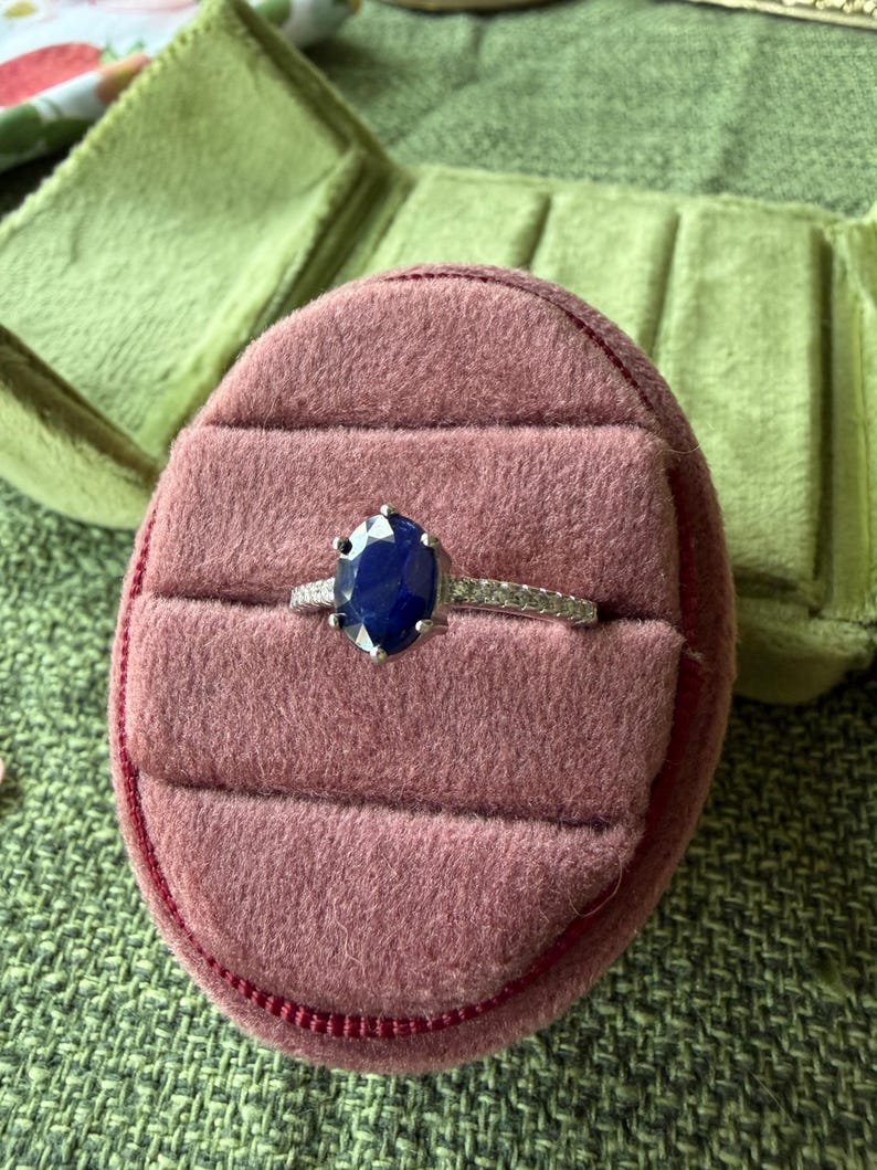 May include: A silver ring with a large, oval, dark blue gemstone, set in a pink velvet ring box. The ring box is inside a green, velvet jewelry case, resting on a green textured surface.