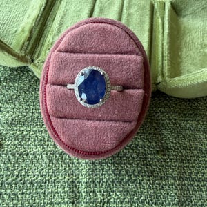 May include: A silver ring with a large, oval, dark blue gemstone, surrounded by a halo of smaller, clear stones. The ring rests in a pink velvet ring box, set against a green textured background.