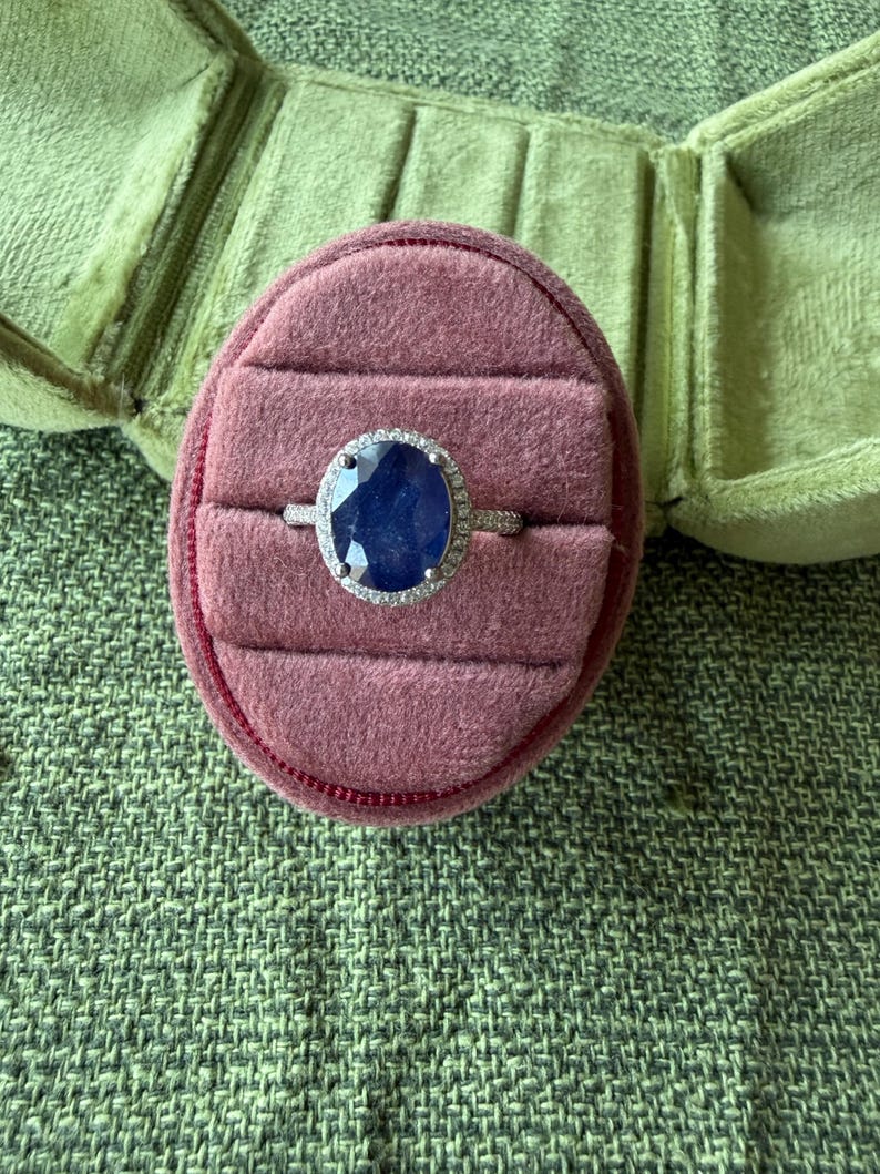 May include: A silver ring with a large, oval, dark blue gemstone, surrounded by a halo of smaller, clear stones. The ring rests in a pink velvet ring box, set against a green textured background.