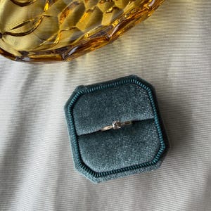 May include: A gold ring with a small, clear gemstone sits in a teal velvet ring box. A vintage amber glass dish is in the background, resting on a light gray fabric surface. The ring box is octagonal.