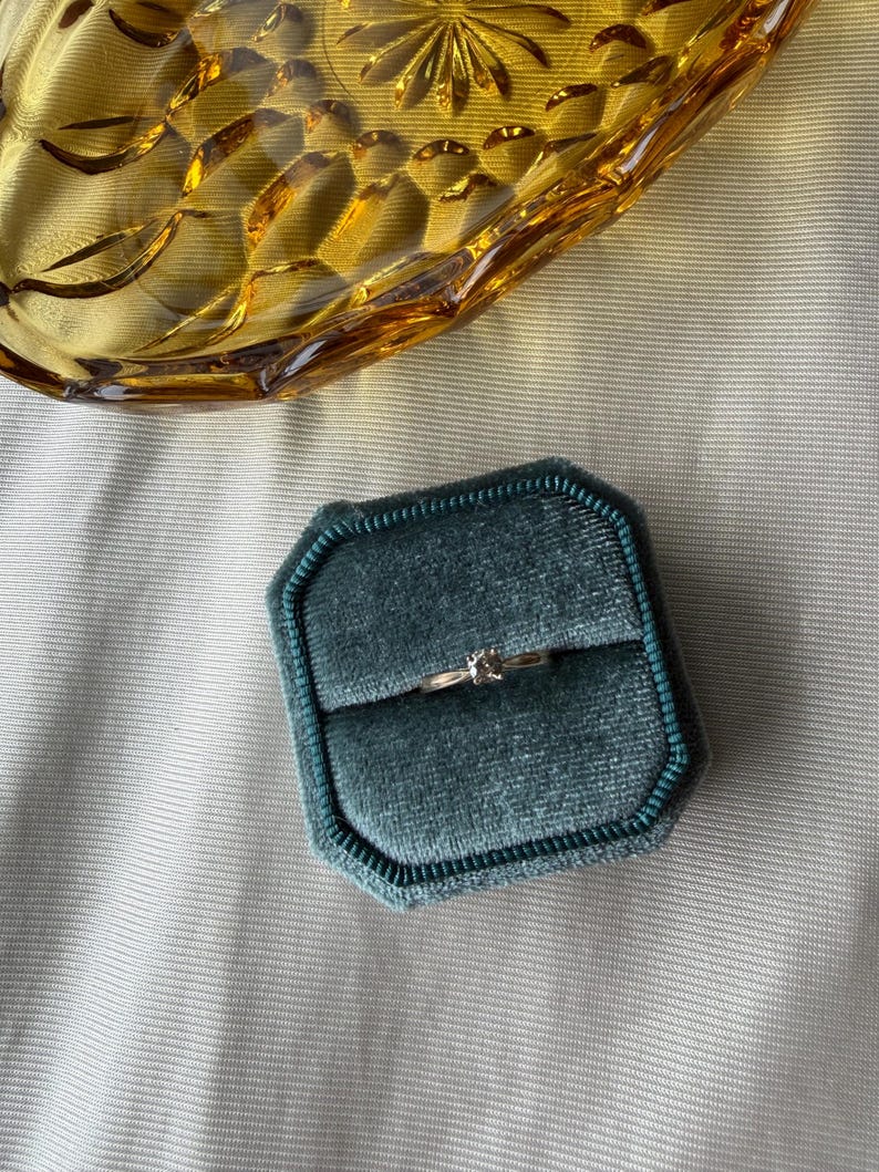 May include: A gold ring with a small, clear gemstone sits in a teal velvet ring box. A vintage amber glass dish is in the background, resting on a light gray fabric surface. The ring box is octagonal.
