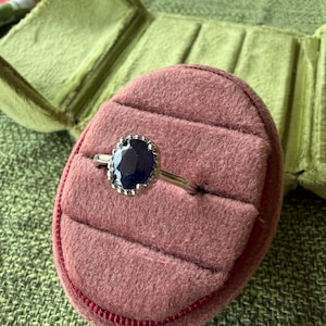 May include: A silver ring with a large, oval, dark blue gemstone. The gemstone is surrounded by a decorative silver setting. The ring is displayed in a pink velvet ring box with a green velvet interior.