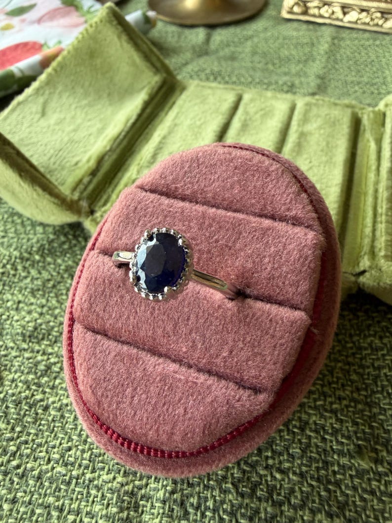 May include: A silver ring with a large, oval, dark blue gemstone. The gemstone is surrounded by a decorative silver setting. The ring is displayed in a pink velvet ring box with a green velvet interior.