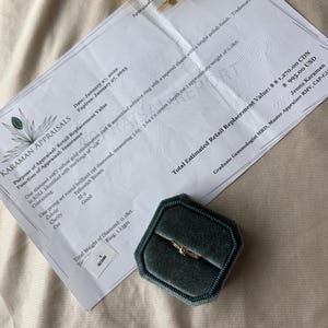 May include: A gold solitaire ring with a diamond, displayed in a dark green velvet ring box. The ring is on a white appraisal document with text. The document includes details about the ring's specifications and value.