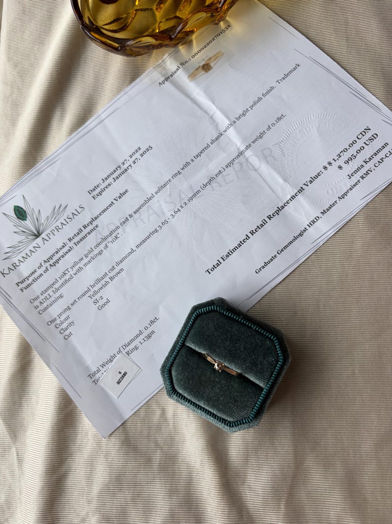 May include: A gold solitaire ring with a diamond, displayed in a dark green velvet ring box. The ring is on a white appraisal document with text. The document includes details about the ring's specifications and value.