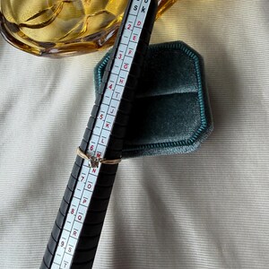 May include: A gold ring with a small diamond is placed on a black ring sizer. The sizer has white markings and numbers. A teal velvet ring box is in the background. The ring size is approximately a size 6.