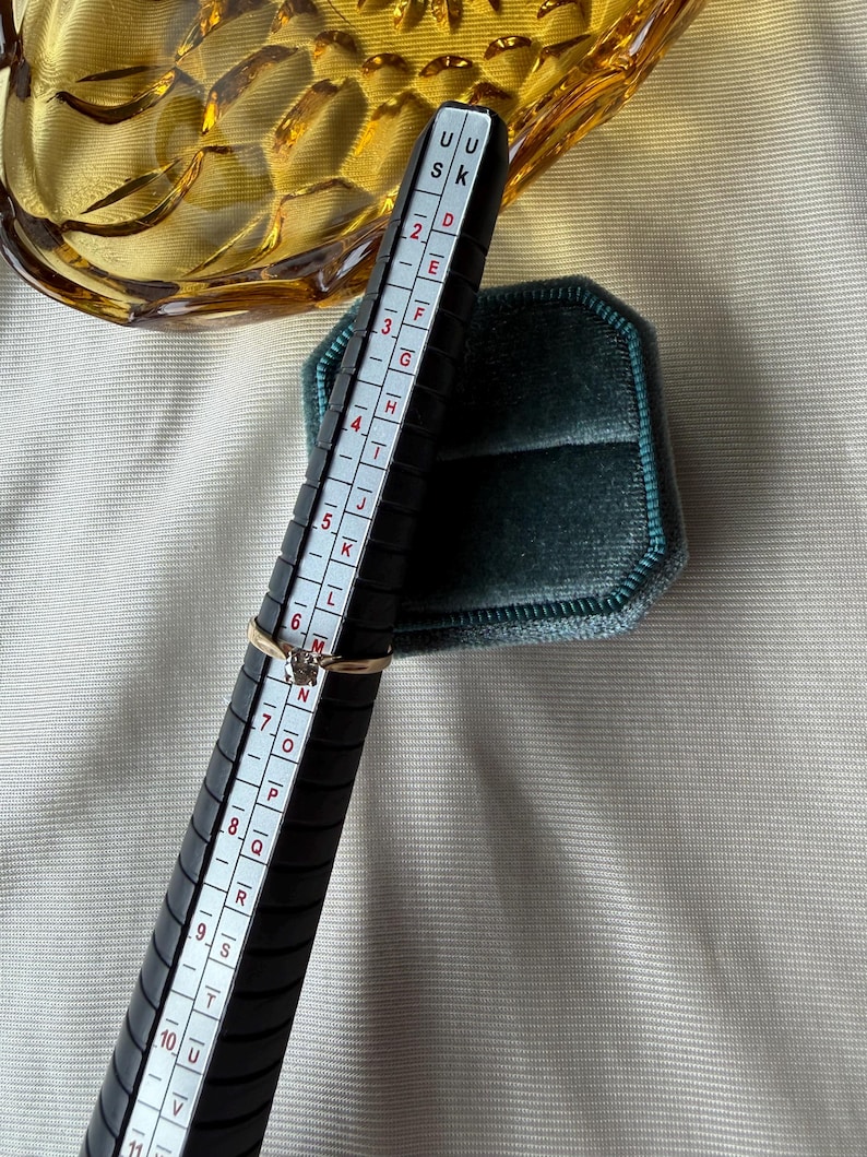 May include: A gold ring with a small diamond is placed on a black ring sizer. The sizer has white markings and numbers. A teal velvet ring box is in the background. The ring size is approximately a size 6.