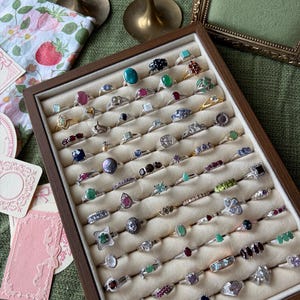 May include: A display case filled with rings of various designs. The rings feature gemstones in colours such as emerald green, ruby red, and sapphire blue. The rings are set in silver and gold bands, displayed in a wooden case.