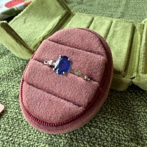 May include: A silver ring with a large, oval, deep blue gemstone is displayed in a pink velvet ring box. The ring box is open, revealing a plush interior. The ring is accented with smaller, clear stones.