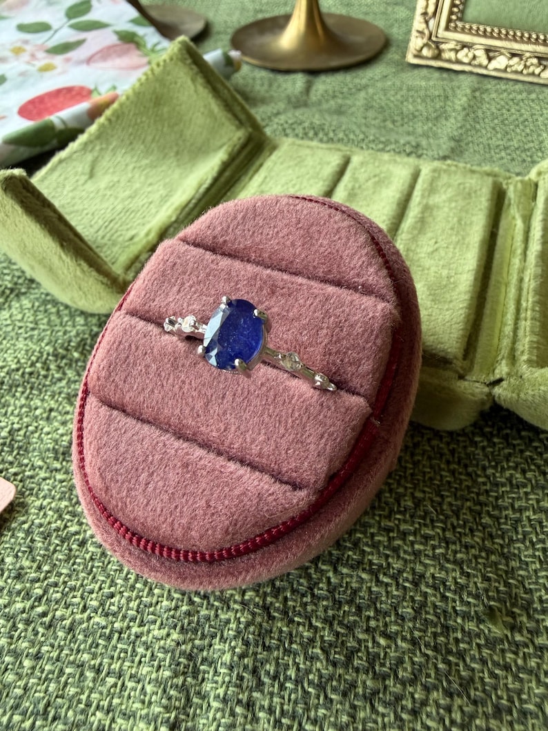 May include: A silver ring with a large, oval, deep blue gemstone is displayed in a pink velvet ring box. The ring box is open, revealing a plush interior. The ring is accented with smaller, clear stones.