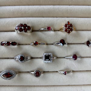 Könnte beinhalten: Eine Sammlung von Edelsteinringen, die in einer Schmuckschatulle präsentiert werden. Die Ringe weisen verschiedene Designs auf, darunter Cluster, Solitäre und Bänder, mit roten und schwarzen Edelsteinen. Die Fassungen sind aus Silber und Gold.