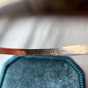 May include: Close-up of a gold-colored bracelet with the letters "9K" stamped on the inside. The bracelet is set against a teal velvet background, with a blurred, golden-toned object in the background.