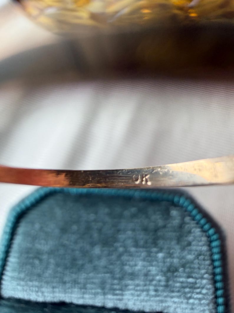 May include: Close-up of a gold-colored bracelet with the letters "9K" stamped on the inside. The bracelet is set against a teal velvet background, with a blurred, golden-toned object in the background.