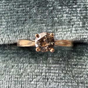 May include: A gold ring with a round, light brown diamond set in a four-prong setting. The ring is set against a teal velvet background, highlighting the ring's design and color.