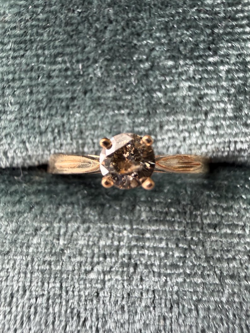 May include: A gold ring with a round, light brown diamond set in a four-prong setting. The ring is set against a teal velvet background, highlighting the ring's design and color.