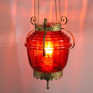 May include: A hanging red glass lantern with a brass frame and chain. The lantern has a ribbed design and a decorative brass base. The light inside the lantern is illuminated, casting a warm glow. The lantern is suspended from a brass fixture.