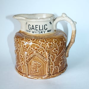 Peut inclure: Pichet en céramique vintage marron et crème. Il présente une anse en forme de branche d'arbre et des motifs floraux et architecturaux en relief. Le texte "GAELIC OLDEST WHISKY BEST" est imprimé sur le dessus. Un profil de femme est en relief sur le devant.