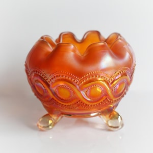 May include: An iridescent orange glass bowl with a scalloped rim and three small feet. The bowl features a detailed pattern of circles and lines around the exterior. The glass has a shiny, reflective quality.