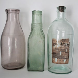 May include: Three glass bottles of varying shapes and colours. On the left, a smooth, light purple bottle with a wide neck. In the centre, a rectangular, light green bottle with a narrow neck. On the right, a clear bottle with a cork and a label.