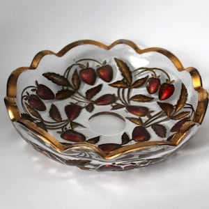 May include: A clear glass bowl with a scalloped edge and gold trim. The bowl is decorated with red strawberries and brown leaves. The bowl has a circular indentation in the center.