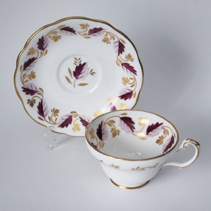May include: A white teacup and saucer with a gold rim and a purple and gold floral design.