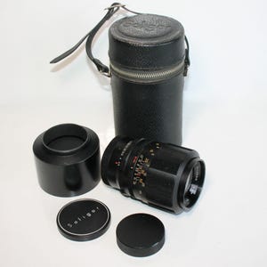 May include: A black Soligor camera lens with a lens hood, lens cap, and a black leather case. The lens has measurement markings and a 135mm focal length. The case has a zipper closure and a handle.