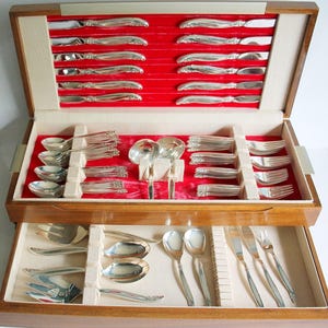 May include: A wooden cutlery chest with two levels, open to show a complete set of polished silverware. The interior is lined with red velvet and beige compartments. The silverware includes knives, forks, spoons, and serving utensils.