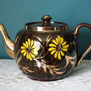 Pode incluir: Uma chaleira marrom com uma borda e uma alça douradas. A chaleira é decorada com flores amarelas e detalhes em folha de ouro.